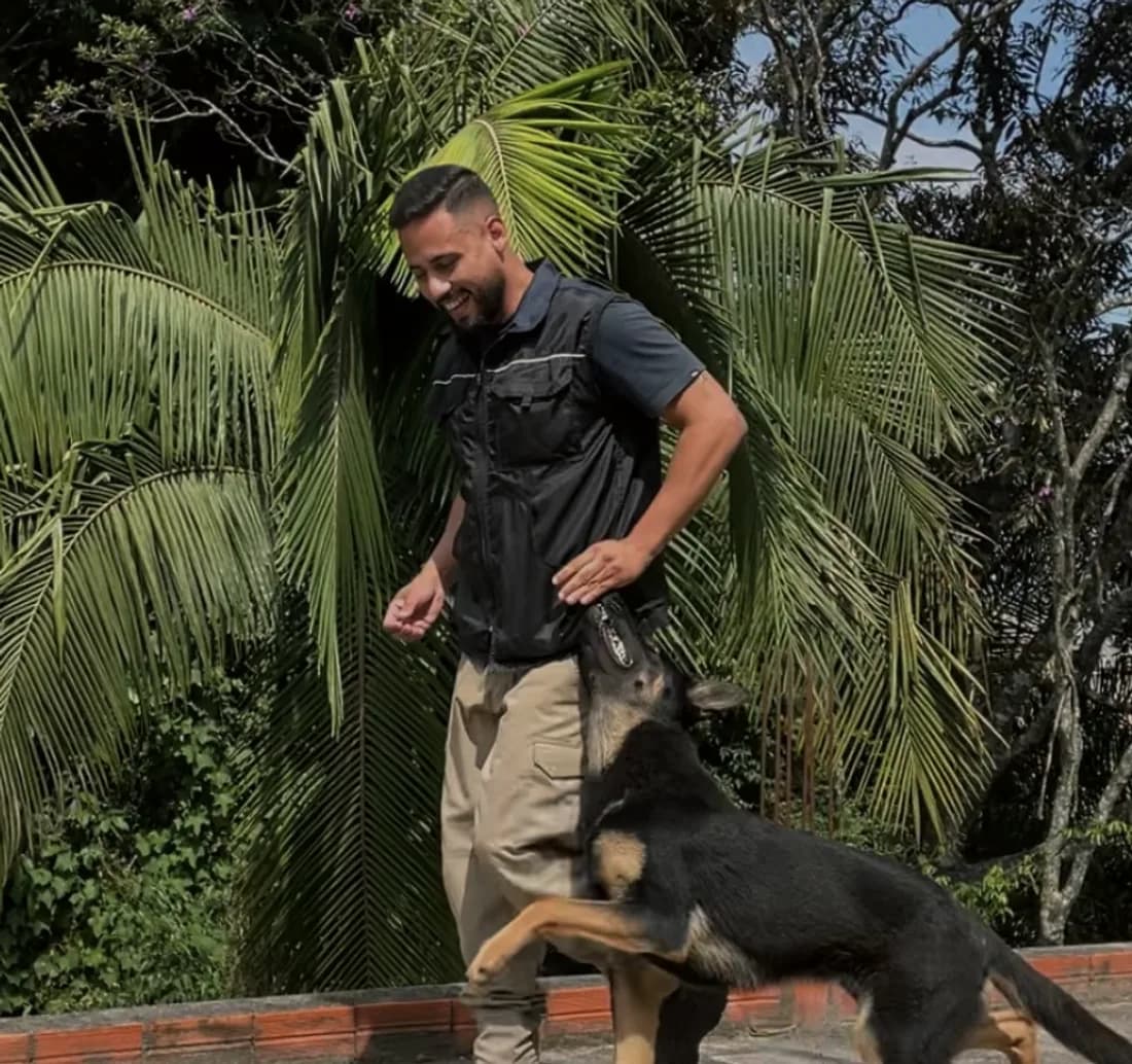 César Leite em pé de colete preto, sorrindo, com um pastor alemão apoiando as patas nele